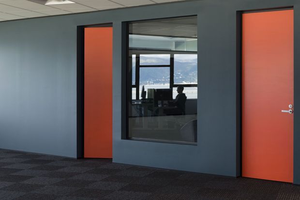 Interior view of a modern commercial office corridor with dark blue-grey walls, bright orange doors in black frames, a large internal observation window, suspended ceiling tiles, and dark patterned carpet. An office workspace and seated person are visible through the glazing, with a mountainous landscape beyond the windows.