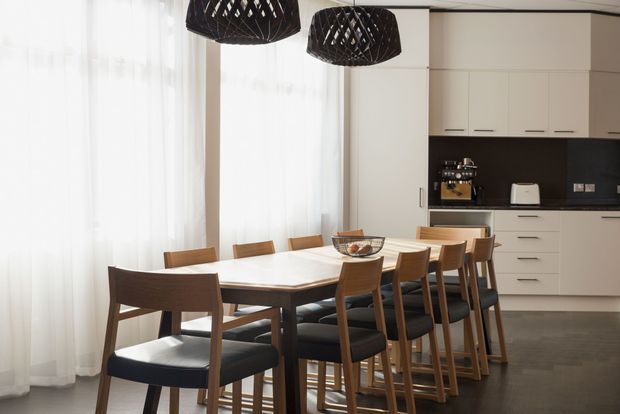 Bright contemporary dining area with a long timber dining table surrounded by mixed dining chairs and stools, positioned beside a minimalist white kitchen. Floor-to-ceiling sheer curtains diffuse abundant natural light, while two black woven pendant lights provide contrast above the table. The kitchen features flat-panel white cabinetry, a dark backsplash and benchtop, and integrated appliances.