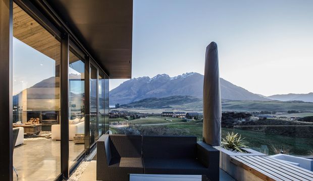 A contemporary covered outdoor terrace or balcony overlooking a dramatic mountain valley and lake or river landscape. The space features dark-framed floor-to-ceiling glazing, a timber-lined ceiling, built-in dark outdoor seating, a sculptural vertical column, and light timber decking or bench surfaces. The interior living area is visible through the glazing, while the primary focus is the elevated outdoor space and expansive alpine outlook.