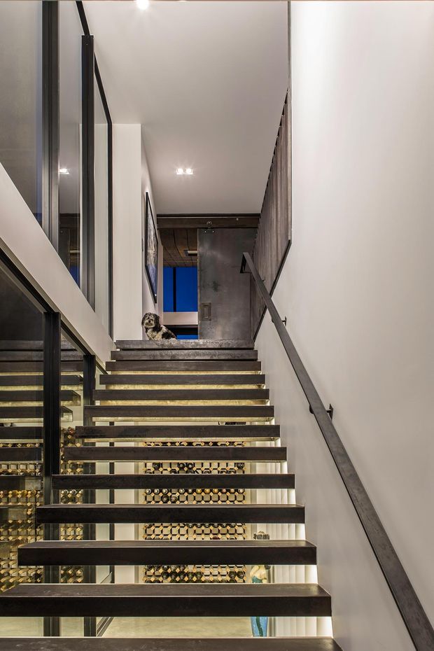 Interior view looking up a modern staircase with open dark metal treads, illuminated riser edges, and a dark metal handrail against white walls. Extensive glazing and reflective surfaces reveal a patterned wine storage area beneath the stairs, with an upper landing and blue-accented space beyond. The composition is architectural and contemporary, with strong vertical lines and layered transparency.