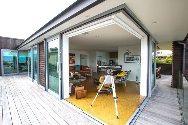 A modern single-level home opening onto a broad timber deck through fully retractable glazed doors. The interior is an open-plan living and dining area with mid-century-inspired furniture, a kitchen visible at the rear, polished warm-toned flooring, white walls, timber wall cladding, and generous indoor-outdoor connection. The deck wraps around the room, creating a bright contemporary entertaining space.