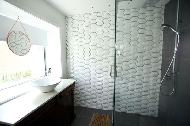 Bright contemporary bathroom with a floating white vessel basin on a dark timber vanity, a large window, and a frameless glass shower enclosure. The shower walls feature white hexagonal tiles with dark grout, contrasted by dark grey wall and floor tiles. A handheld shower is mounted on the right wall, and a small timber bath mat adds warmth.