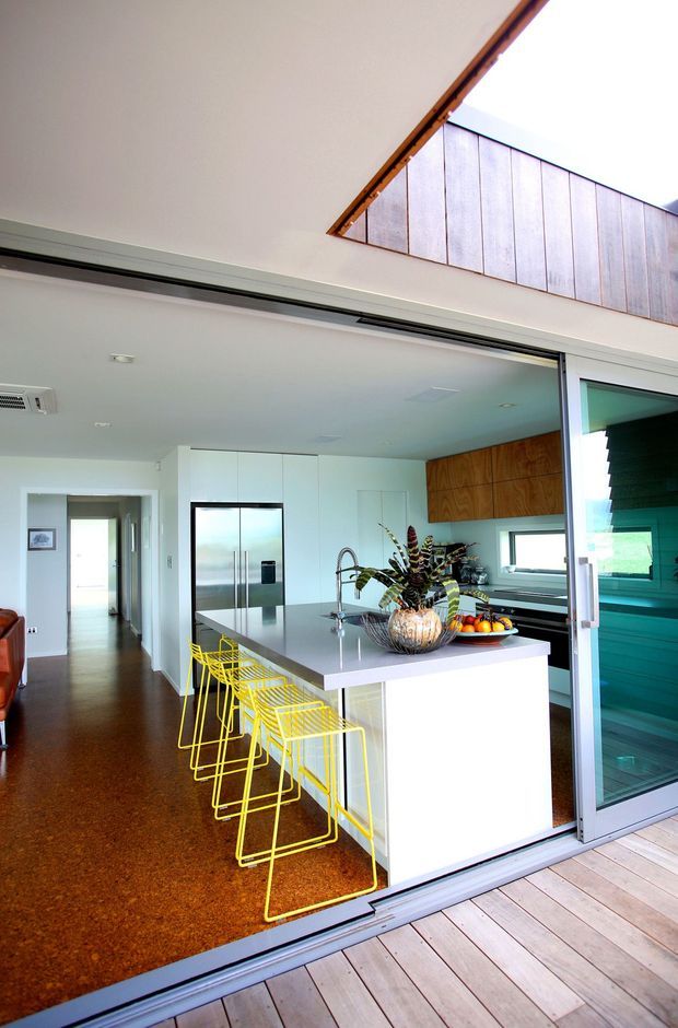 Bright contemporary kitchen viewed from a timber deck through wide sliding glass doors. The open-plan kitchen features a large white island with a pale stone benchtop, integrated white cabinetry, stainless-steel refrigerator and appliances, a timber feature wall of cabinetry, and vivid yellow wire bar stools. A hallway extends beyond the kitchen, while a raised glazed clerestory or upper window adds natural light.