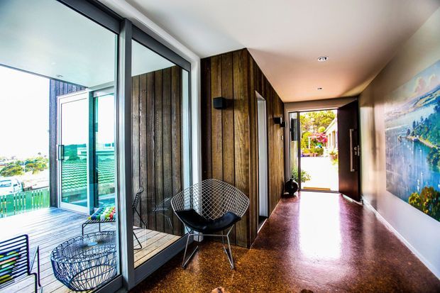 A bright contemporary residential hallway with a polished dark floor, vertical timber feature cladding, full-height glazed doors and windows opening to a deck, and a black wire-frame lounge chair. A large colourful artwork lines the right wall, while an open entry door at the far end connects to the outdoors.