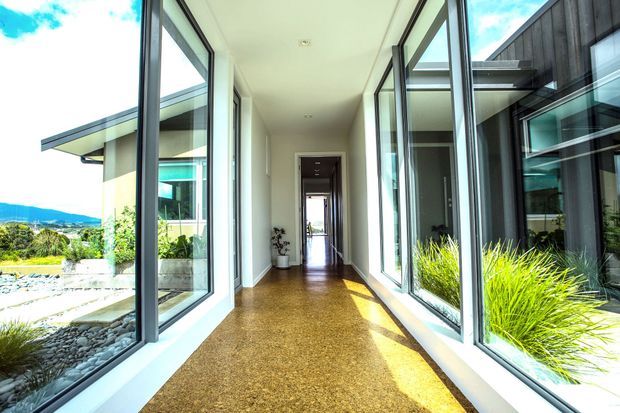 A bright, glazed architectural corridor or gallery-like passageway with floor-to-ceiling windows on both sides. The corridor has a polished warm brown concrete or terrazzo-like floor, white walls and ceiling, dark slim window frames, and views onto landscaped courtyards with ornamental grasses and stone. A darker timber-clad building volume is visible through the glazing, with distant hills beyond.