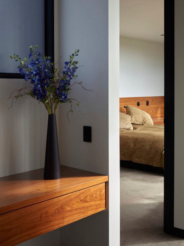 A refined interior view of a minimalist entryway or hallway, featuring a floating dark timber console with a richly grained timber front, a tall matte-black vase with blue flowers, and a black wall switch. Through the doorway, a softly styled bedroom is visible with a low bed, beige bedding, timber wall panelling, and carpeted flooring. The composition uses strong vertical lines, recessed lighting, and a restrained neutral palette with warm timber accents.
