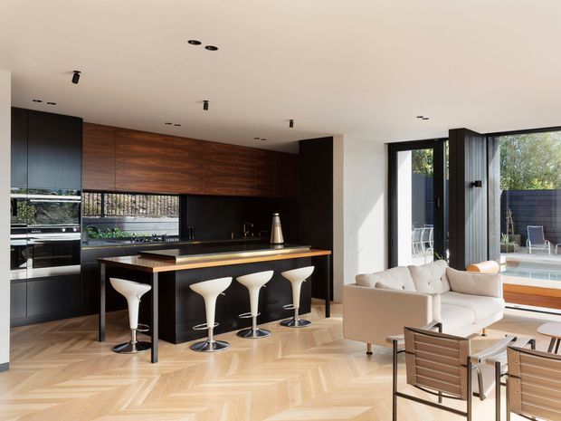 A bright, open-plan contemporary kitchen and living area. The kitchen features dark matte cabinetry, a warm timber wall and island front, a dark benchtop, integrated appliances, and four white sculptural bar stools. The room opens to a living area with pale upholstered seating and large black-framed glazing overlooking an outdoor pool area. A light herringbone timber floor and recessed ceiling lighting complete the refined interior.