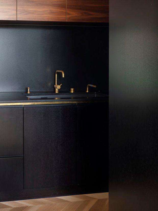 A compact, dramatically dark kitchen featuring black textured cabinetry, a dark benchtop and splashback, warm brass or bronze tapware and trim, and rich walnut overhead cabinetry. A light and mid-toned timber herringbone floor provides contrast at the base of the image.
