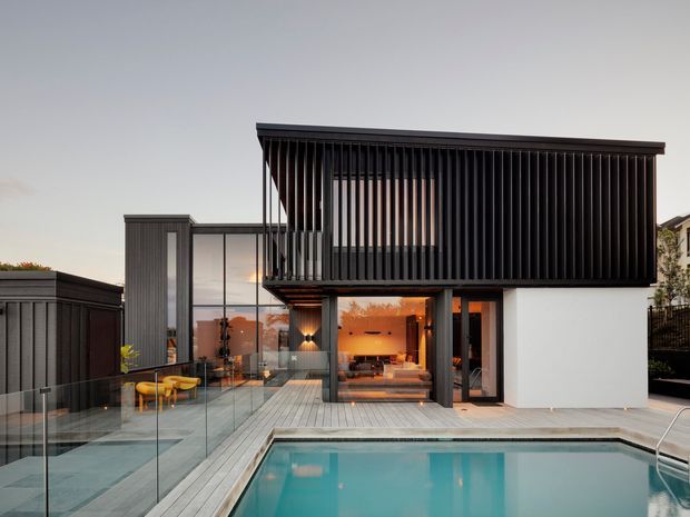 Contemporary multi-level residence viewed from the pool deck at dusk. The home features a crisp white lower volume, extensive glazing, dark vertical external screening on the upper level, and a timber deck surrounding a rectangular swimming pool. A glass balustrade encloses an elevated terrace, with warm interior lighting visible through the open living areas.