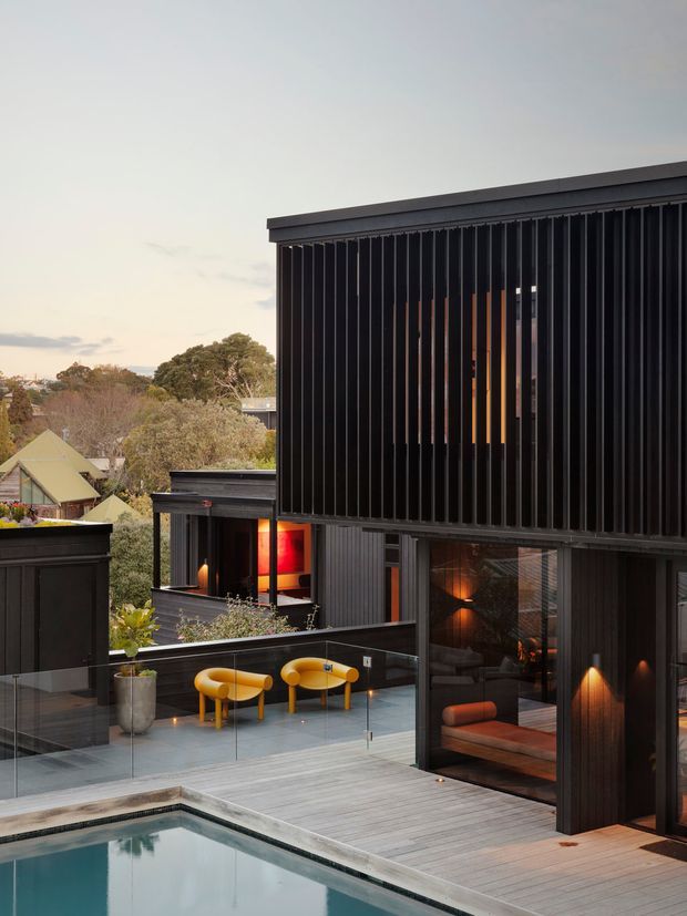 Exterior view of a contemporary multi-level dark-clad residence with strong vertical screening, large glazed openings, and a rooftop or elevated timber deck beside a swimming pool. The architecture features black vertical battens and metal-like cladding, warm interior lighting, glass balustrades, and a compact outdoor seating area with bright yellow chairs. Suburban houses and trees are visible in the background.