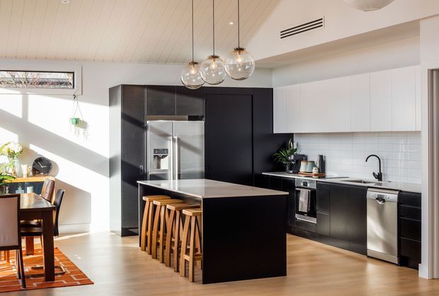 Bright contemporary kitchen with a large black island, integrated dark cabinetry, white upper cabinets, stainless-steel appliances, a white tiled splashback, and timber bar stools. The open-plan space also includes a small dining area, warm timber flooring, a light timber-lined ceiling, and globe pendant lighting.