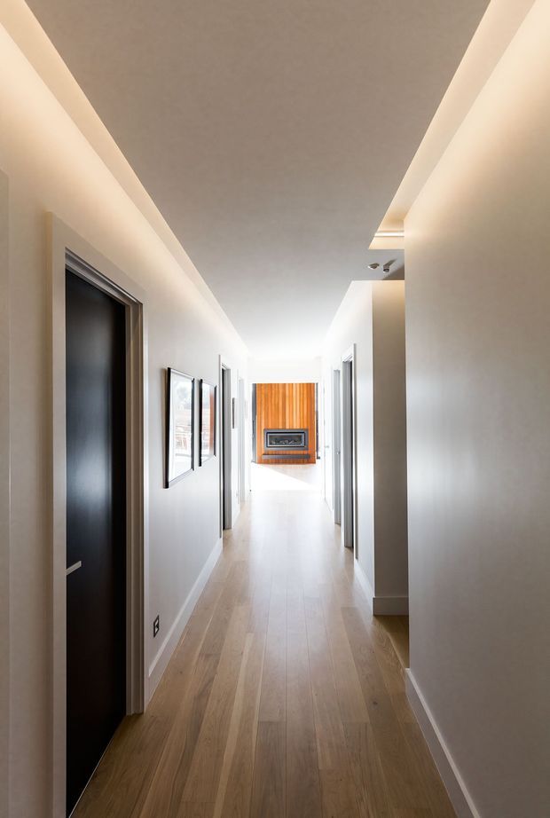 A long, bright residential hallway with light timber flooring, smooth white walls, recessed cove lighting, multiple dark-framed internal doors, framed artwork, and a timber feature wall with a built-in fireplace or display at the far end. The space has a clean, contemporary architectural character.