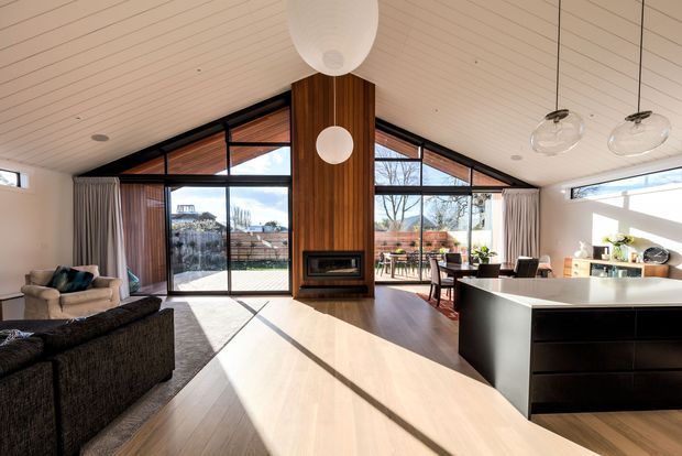 Open-plan living, dining and kitchen interior with a dramatic pitched timber-lined ceiling and extensive black-framed glazing. A central vertical timber feature wall incorporates a built-in fireplace, while the pale timber floor, dark kitchen island, contemporary pendant lights and neutral furnishings create a warm, modern space. The glazing opens to an outdoor entertaining area and garden beyond.