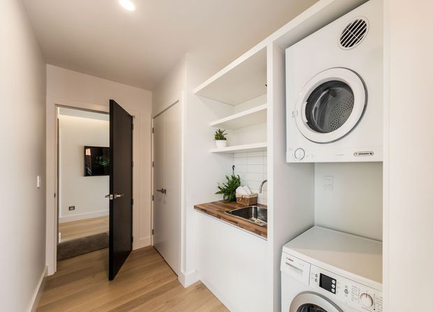 Bright, compact laundry and utility area set within a hallway. A stacked white washer and dryer occupy a built-in alcove, while adjacent white cabinetry includes open shelving, a timber benchtop, stainless-steel sink, and white tiled splashback. Light timber flooring and warm white walls create a clean, practical interior.