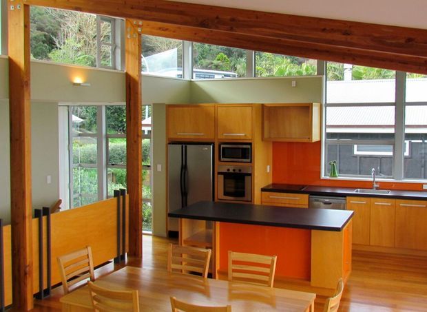 Open-plan kitchen and dining area with extensive warm timber structure and flooring, pale green walls, and large windows. The kitchen features warm wood cabinetry, a dark island benchtop, stainless-steel appliances, and a long window-side countertop. A timber dining table and chairs occupy the foreground, while exposed timber beams and posts create a strong structural presence overhead and along the sides.