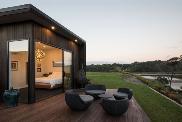 A compact modern pavilion or retreat with dark vertical timber cladding, a flat roof and extensive corner glazing, opening onto a timber deck furnished with rounded black outdoor lounge chairs and a low table. The interior bedroom is visible through the glass, while the deck overlooks a landscaped grassy setting and a calm waterway at dusk.