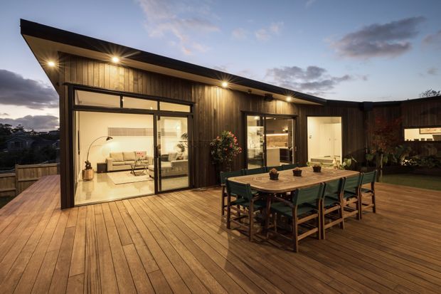A single-storey contemporary home viewed from a large timber deck at dusk. The dark vertically clad facade has a dramatic asymmetrical pitched roof with deep eaves, extensive sliding glass doors, warm exterior lighting, and an outdoor dining setting. A brightly lit living room is visible through the glazing, while fencing and neighbouring homes appear in the background.