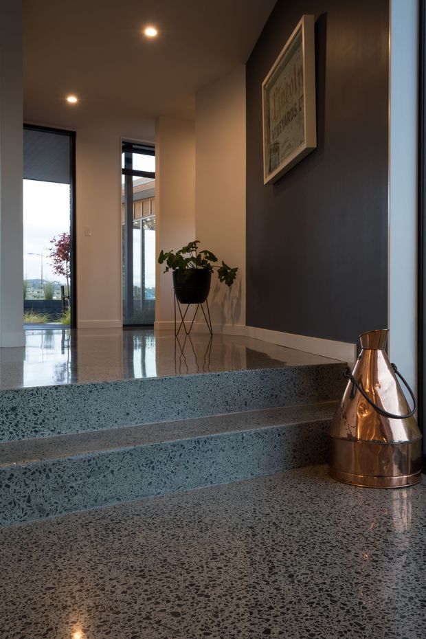 Interior view of a refined entry hall with broad polished terrazzo steps and flooring, dark charcoal feature wall, warm recessed lighting, large glazed doors, a potted plant, and framed artwork. A copper-toned decorative vessel sits beside the stairs.