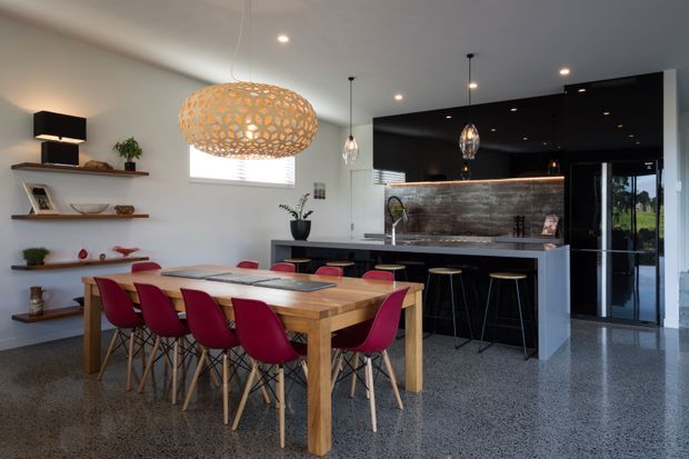 Open-plan kitchen and dining area with a substantial timber dining table surrounded by red upholstered chairs, floating timber display shelves, pendant lighting, and a dark contemporary kitchen with a large island and metallic textured splashback. Polished concrete or terrazzo-like flooring and neutral walls create a refined modern interior.