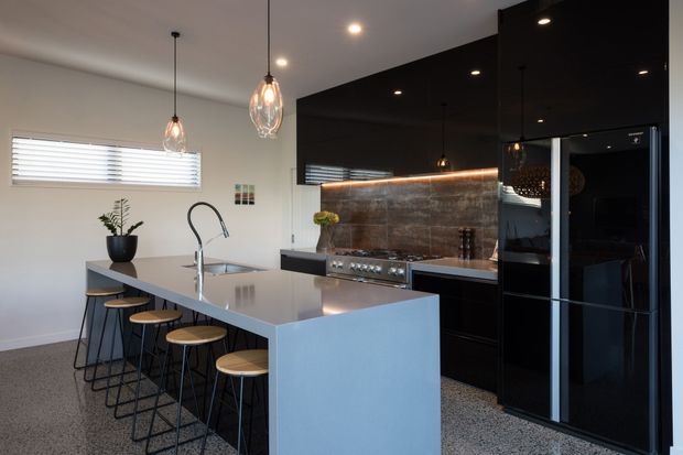 Contemporary kitchen featuring a large pale grey island with integrated sink and seating for four, glossy black cabinetry, a dark metallic textured splashback, stainless-steel cooktop, black appliances, pendant lights, and polished concrete-style flooring. The space is compact-to-medium in size with a clean, high-contrast palette.