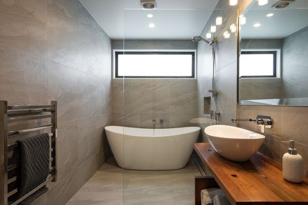 Contemporary bathroom with a freestanding white bathtub, frameless glass shower enclosure, vessel sink on a timber vanity, large wall mirror, heated towel rail, and warm grey stone-look tiled walls and floor. A high horizontal window provides natural light.