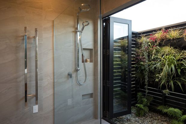 Contemporary walk-in shower with large-format light stone-look wall tiles, exposed chrome shower fittings, and a frameless glass screen. An open black-framed door connects directly to a small landscaped courtyard featuring a vertical garden, black horizontal cladding, gravel, and lush planting.