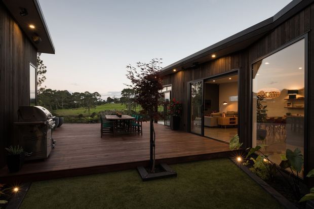 A contemporary single-storey home arranged around a landscaped courtyard, viewed from an outdoor timber deck. Dark vertical timber cladding, broad glazed sliding doors, warm interior lighting, outdoor dining, built-in barbecue, and a low-pitched roof create a cohesive indoor-outdoor entertaining space. The surrounding rural greenery is visible beyond the courtyard.