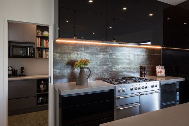 A sophisticated dark-toned kitchen featuring a large stainless-steel range cooker, black built-in cabinetry, a mottled metallic-looking splashback, and a dark glossy ceiling with recessed and pendant lighting. An open pantry or compact utility storage area with shelving, microwave, appliances, and books is visible on the left. A peninsula or benchtop occupies the foreground.