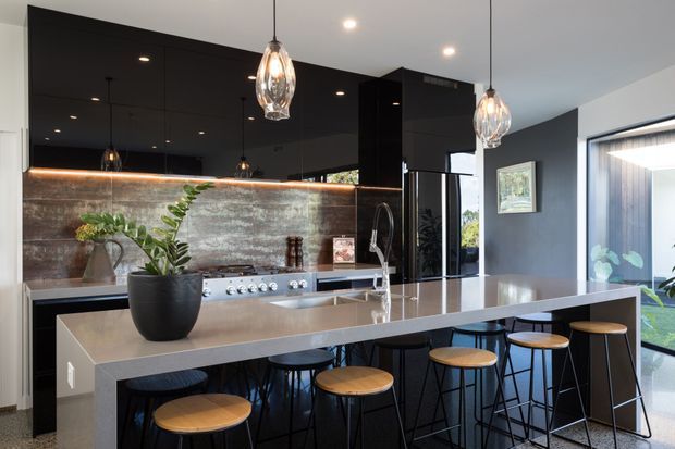 A sophisticated contemporary kitchen with a large stone island and integrated breakfast bar, black high-gloss cabinetry, a textured metallic-look splashback, and a stainless steel cooking zone. Pendant lights hang above the island, while timber-topped stools provide casual seating. Large glazing and a glazed door bring in natural light, with plants adding a small organic accent.