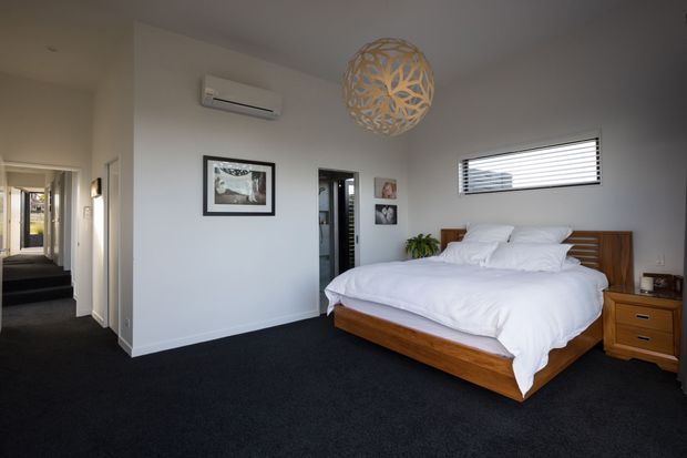 A clean, contemporary bedroom with a low-profile timber bed and matching bedside table, crisp white bedding, dark carpet, white walls, a horizontal louvre window, framed artwork, and a sculptural pendant light. A hallway and adjoining spaces are visible through the doorway.
