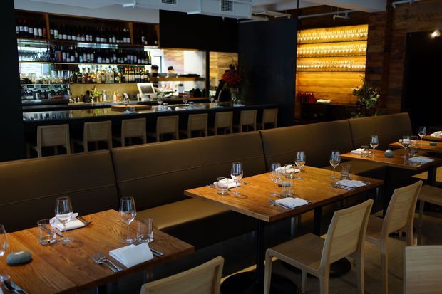 A warmly lit restaurant interior with a prominent wood bar and backbar shelving, illuminated bottle displays, a long upholstered banquette, and multiple timber dining tables set with glassware and table settings. The space has dark timber finishes, exposed ceiling services, and a refined hospitality atmosphere.