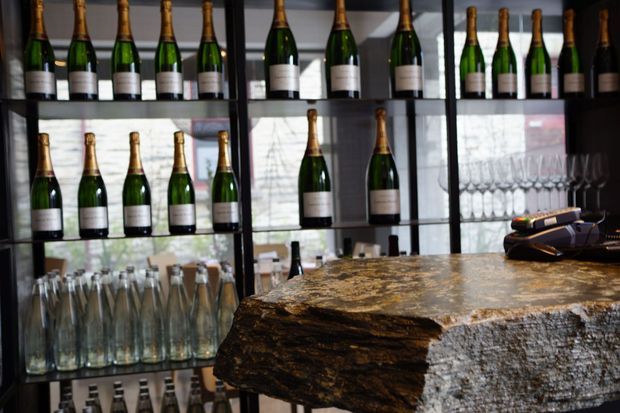 A stylish bar interior featuring dark-framed glass shelving filled with champagne bottles and glassware. A substantial rough-edged natural stone or timber-look counter dominates the foreground, with a bar till and additional bottles and glasses visible behind it. The space has a moody, refined hospitality atmosphere with strong indoor-outdoor light through the glazing.