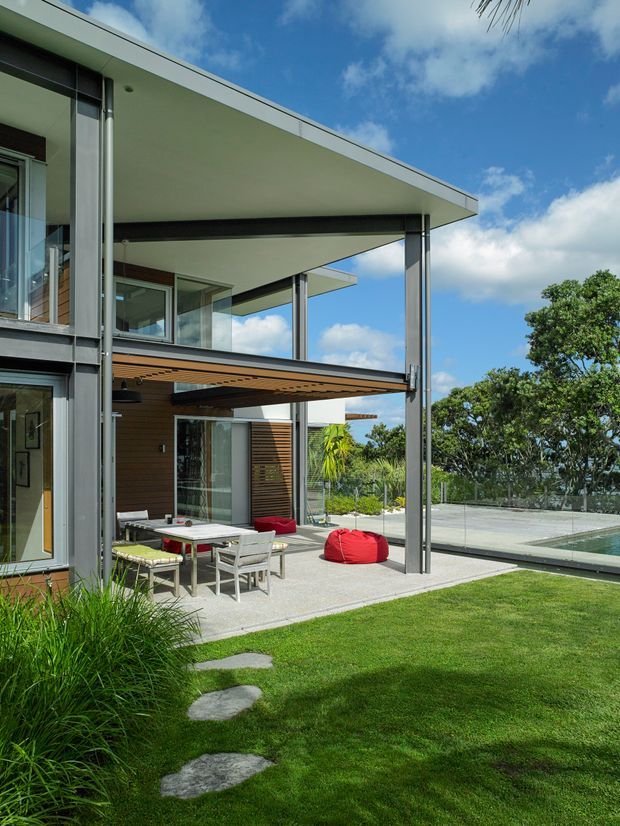 A contemporary multi-level home with broad flat roof planes, extensive floor-to-ceiling glazing, slim steel or aluminium posts, timber-lined soffits, and horizontal timber cladding. The image focuses on a covered outdoor entertaining terrace with a dining setting and red beanbag beside a lawn and swimming pool. Lush planting and a bright blue sky surround the house.