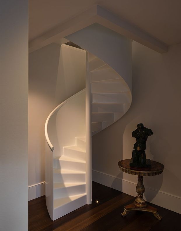 A warmly lit interior view of a sculptural white spiral staircase with a curved balustrade and central supporting form. The staircase rises through a compact stairwell framed by white painted walls and beams, with dark timber flooring below. An ornate round pedestal table with a dark decorative statue occupies the corner beside the stairs.