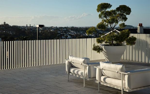 A contemporary rooftop terrace with a ribbed light-grey deck, tall vertical slatted privacy screen, and two white cushioned outdoor armchairs. A large sculptural potted tree anchors the seating area, with a broad suburban cityscape visible beyond the terrace.