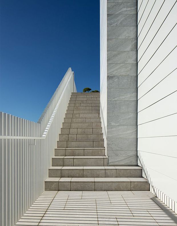 A minimalist exterior stairway rises between a white vertically slatted screen and a white horizontally panelled wall. Large-format grey stone or concrete tiles cover the steps and landing, while a tall grey stone-clad wall forms the central architectural element. Strong linear shadows from the slatted screen create a graphic pattern across the paving in bright sunlight, with a deep blue sky visible above.