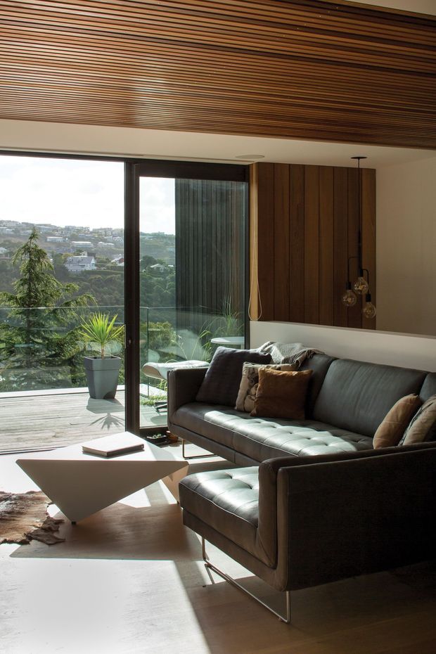 A refined contemporary living room with a large dark upholstered sectional sofa, sculptural white coffee table, timber-paneled feature wall, and slatted timber ceiling. Full-height glazing opens to a timber deck and leafy suburban landscape, bringing in abundant natural light.