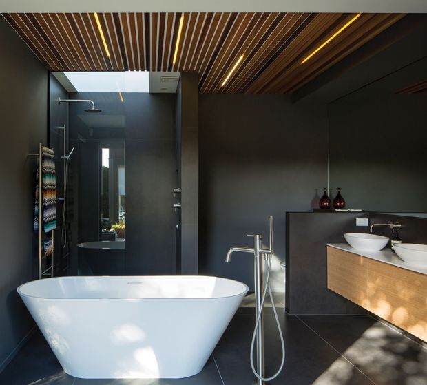 Contemporary dark-toned bathroom featuring a freestanding white bathtub, walk-in glass shower, double vessel vanity, and wall-mounted timber-front cabinetry. The room has large dark floor and wall tiles, a dramatic timber-slat ceiling with integrated linear lighting, and a skylight bringing in natural light.