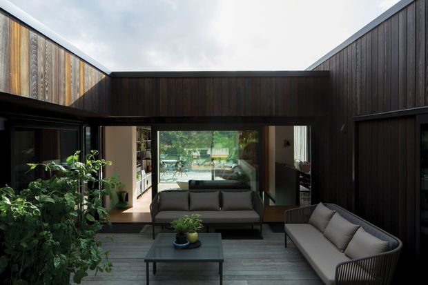 A dark timber-clad courtyard or outdoor lounge framed by tall enclosing walls, with a grey outdoor sofa, coffee table, potted plants, and a glazed opening into the living space. The courtyard creates a sheltered, inward-looking setting with strong indoor–outdoor connection and reflected greenery visible through the glazing.