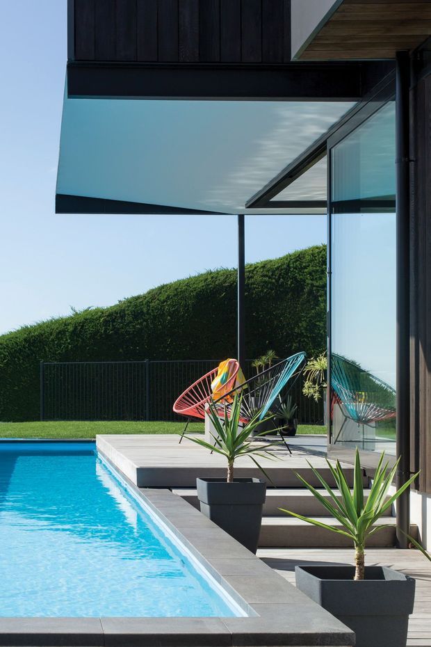 A contemporary modern poolside terrace beside a dark-clad house, featuring a long rectangular swimming pool, pale timber decking, large frameless-looking glazed openings, deep roof overhangs, sculptural potted yucca plants, and colourful wire lounge chairs. The image emphasizes the crisp indoor-outdoor connection and strong geometric lines in a bright landscaped setting.