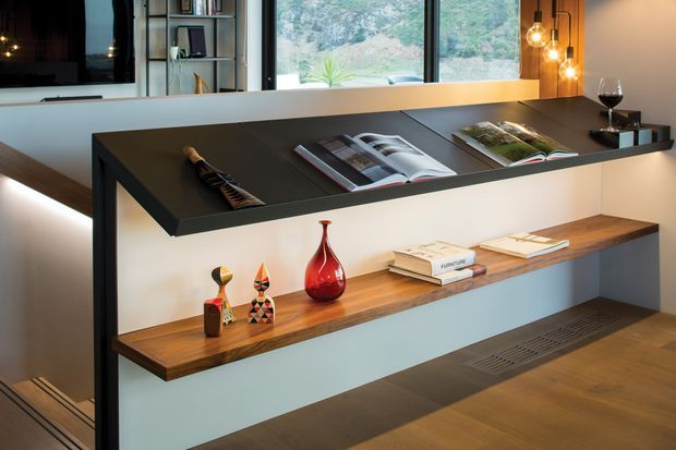 Contemporary built-in media and display joinery with a dark cantilevered upper shelf, warm timber lower shelf, integrated lighting, books, decorative objects, and a wall-mounted television. Large glazing behind the shelving looks onto a wooded rocky landscape, while pendant bulbs add a warm accent.