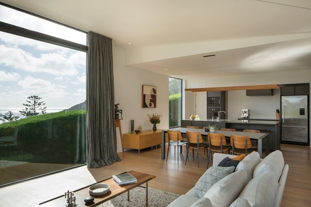 Open-plan contemporary living, dining, and kitchen area with floor-to-ceiling glazing overlooking a coastal landscape. The room features warm timber flooring, a streamlined dark kitchen with island seating, a pale sectional sofa, timber dining furniture, and full-height grey curtains.