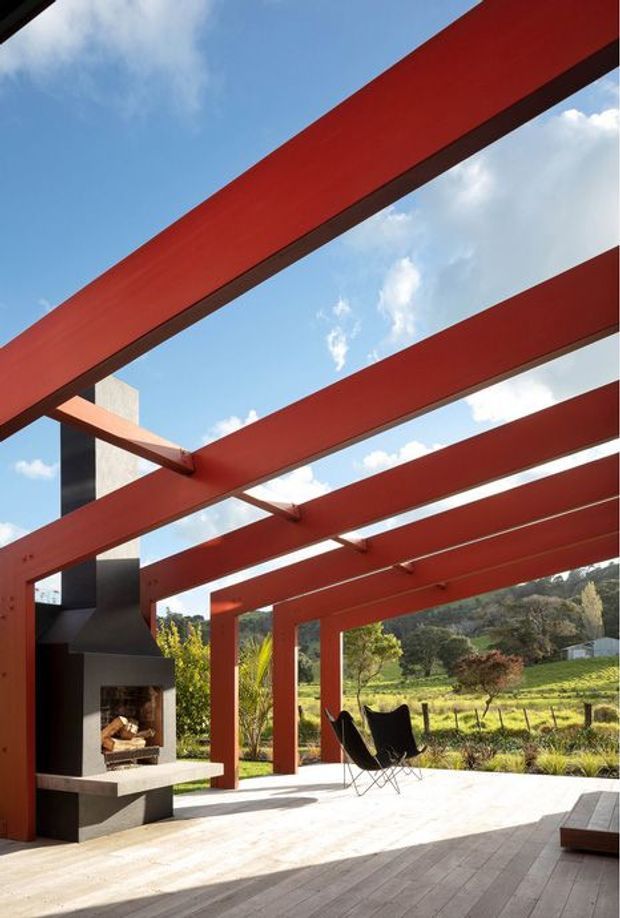A covered outdoor terrace with bold red-painted exposed steel beams and columns, opening to a rural landscaped setting. A built-in dark masonry outdoor fireplace with a projecting concrete hearth anchors the left side, while black sling chairs and a timber-toned deck create a relaxed entertaining area. The open pergola-like roof structure frames a bright blue sky and surrounding hills.