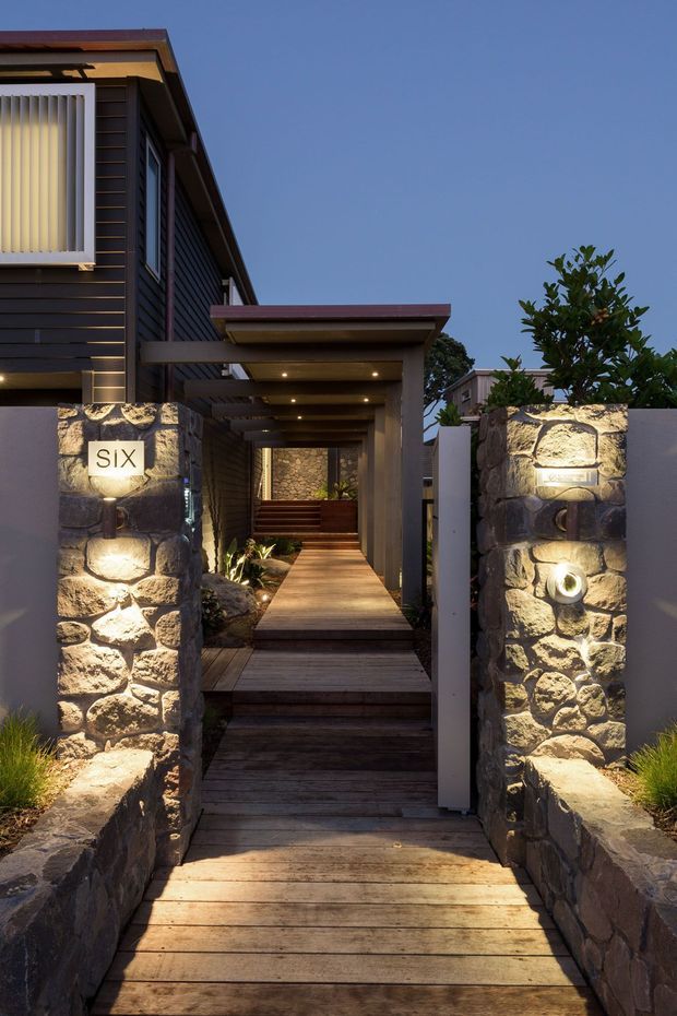 A dusk exterior view of a residential entrance approached by a timber boardwalk. Rough stone gate pillars with illuminated house-number plaques frame the path, leading beneath a covered porch toward a dark-clad, multi-level home. Warm recessed lighting highlights the timber decking, stonework, and planted edges.
