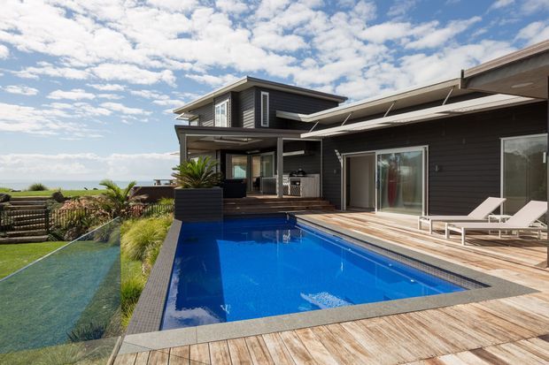 Contemporary multi-level dark-clad residence viewed from its outdoor pool terrace. The scene features a vivid blue swimming pool, timber-look decking, white sun loungers, extensive glazing, covered outdoor areas, and landscaped planting, with an open coastal horizon visible beyond.