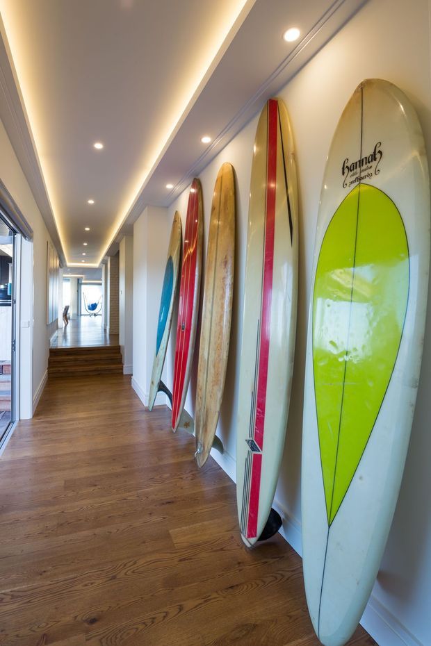 A bright, narrow hallway with warm timber flooring, white walls, recessed ceiling lights, and concealed linear lighting. Several colorful surfboards are displayed upright along the right wall, while glazing on the left brings in natural light. The corridor leads to a stepped area and a bright open space beyond.