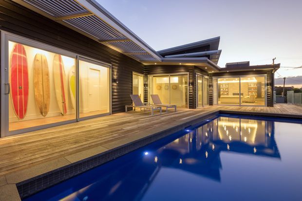 Twilight exterior view of a contemporary single-storey home arranged around a swimming pool. The dark horizontal cladding, angular roof forms, extensive glazing, timber decking, illuminated surfboard display, and outdoor lounge create a polished resort-like setting.