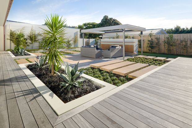 Contemporary landscaped backyard with expansive grey timber decking, geometric raised garden beds, architectural planting, stepping-stone pavers, and a covered outdoor entertaining pavilion furnished with outdoor seating and a bar or kitchen zone.