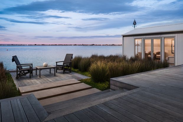 A calm waterfront outdoor living area with expansive dark timber decking, a lower seating platform beside the water, and two lounge chairs around a small table. Ornamental grasses soften the edge of the deck, while a compact light-clad pavilion with full-height glazed doors opens onto the deck. The low pitched metal roof includes a small decorative finial, and the scene is shown at dusk beneath a blue and pink sky.
