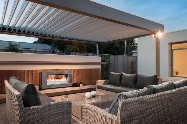 An elegant covered outdoor lounge with woven seating arranged around a low coffee table and a built-in linear fireplace set into a timber-clad feature wall. A large louvred pergola provides overhead shade, while an illuminated interior room is visible through the doorway at right.